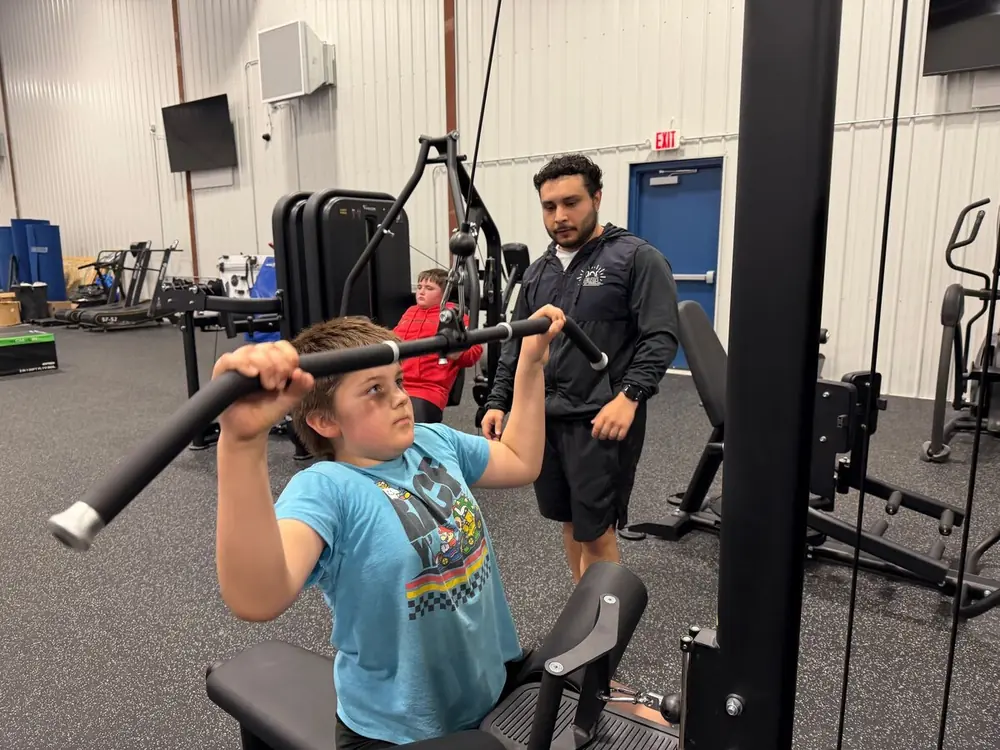 A boy on the lat pull-down machine with a coach guiding his form